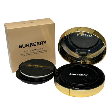 Burberry Ultimate Glow Cushion foundation shown with its gold and black packaging box, open gold compact, and black foundation puff on a white background.