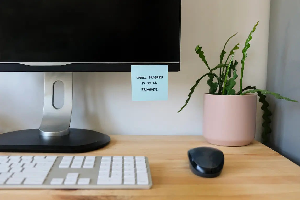 A minimalist desk setup featuring a black computer monitor with a silver stand, a white keyboard, a black mouse, and a small potted green plant in a pink pot. A light blue sticky note is taped to the monitor arm with the handwritten text 'SMALL PROGRESS IS STILL PROGRESS'.