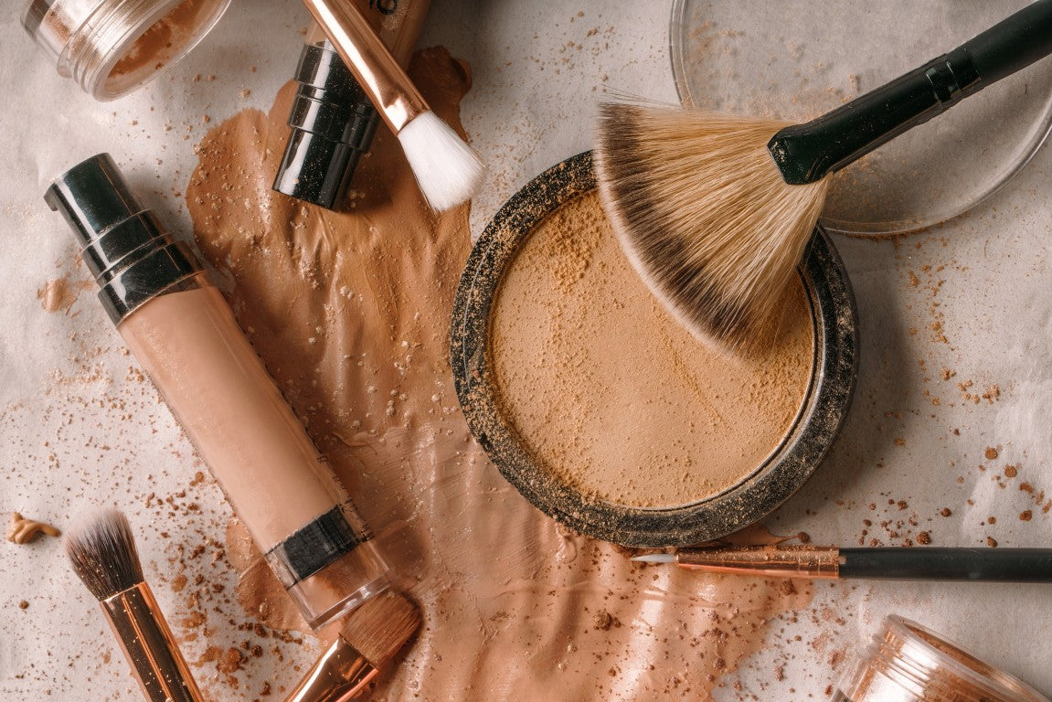 Makeup products including a concealer, brushes, and a bowl of powder on a textured surface.