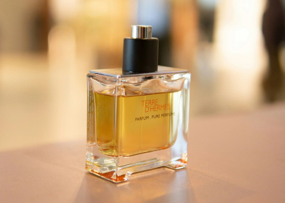 A bottle of Terre d'Hérmités Parfum Pure Persilie perfume, featuring a clear glass bottle filled with golden-yellow liquid, a black cylindrical cap, and a silver top, set against a warm, blurred background.